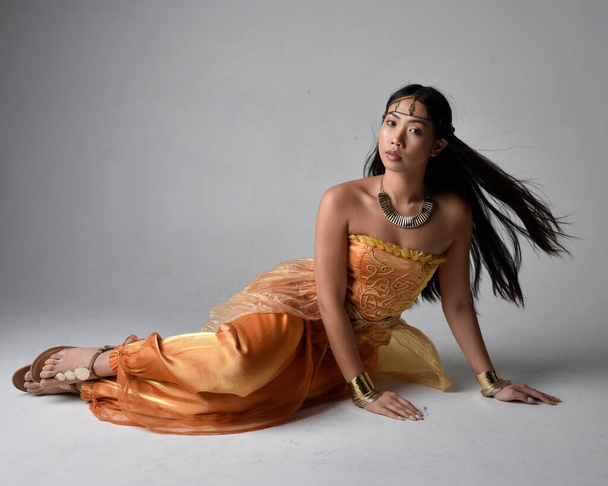 Full length portrait of pretty young asian woman wearing golden Arabian robes like a genie, seated pose, isolated on studio background. - Foto, imagen
