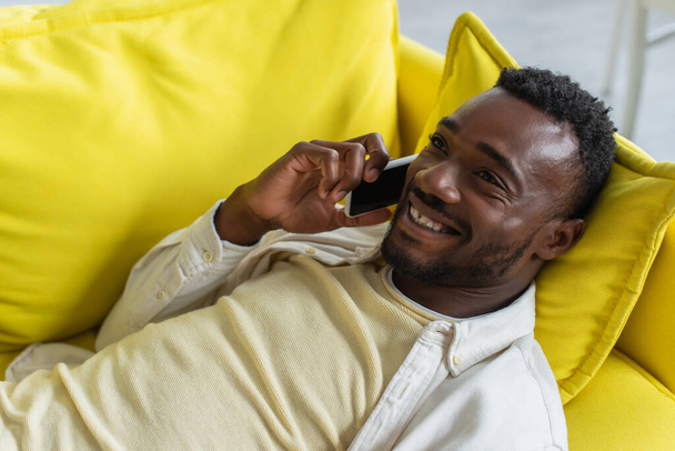 happy african american man talking on smartphone while lying on yellow sofa - Фото, зображення