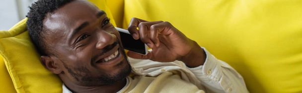 Χαρούμενος Αφροαμερικάνος που μιλάει στο smartphone ενώ ξαπλώνει στον κίτρινο καναπέ, πανό - Φωτογραφία, εικόνα