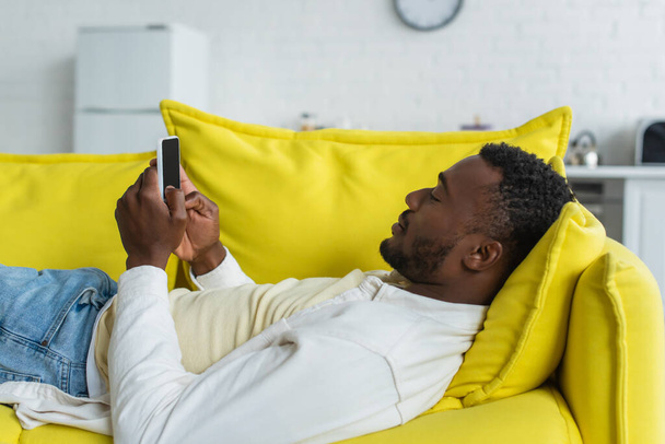 african american man using smartphone while lying on yellow sofa - Fotó, kép