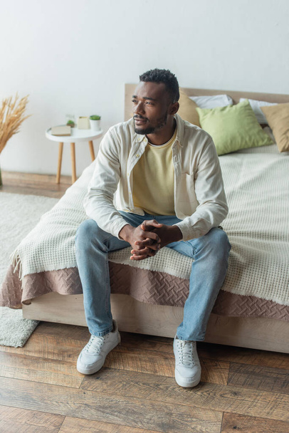 pensativo joven afroamericano hombre con las manos apretadas sentado en la cama - Foto, Imagen