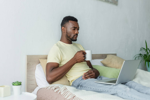 bearded african american freelancer using laptop and holding cup of coffee in bedroom  - Foto, imagen