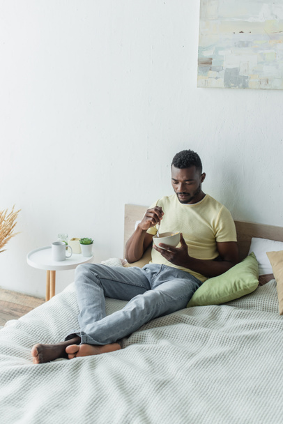 barefoot african american man looking at corn flakes in bowl while chilling in bed - Photo, image