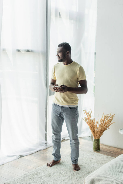 πλήρες μήκος γενειοφόρου Αφροαμερικανού άνδρα στέκεται με smartphone  - Φωτογραφία, εικόνα