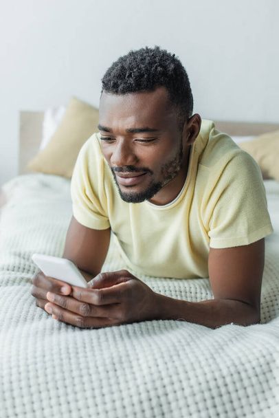 alegre afroamericano hombre en camiseta usando smartphone y acostado en la cama - Foto, Imagen