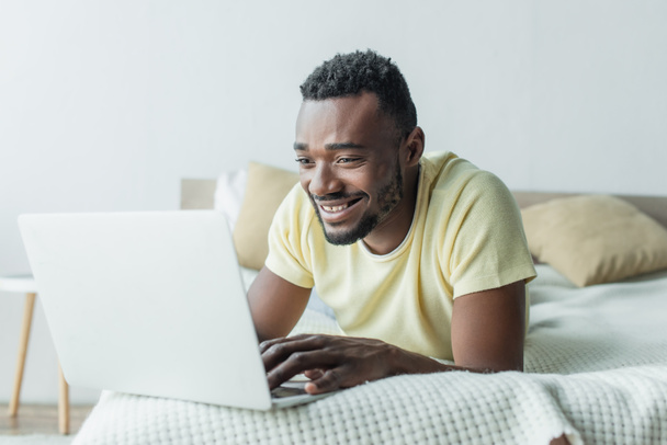veselý afroameričan na volné noze v tričku pomocí notebooku a ležící na posteli - Fotografie, Obrázek