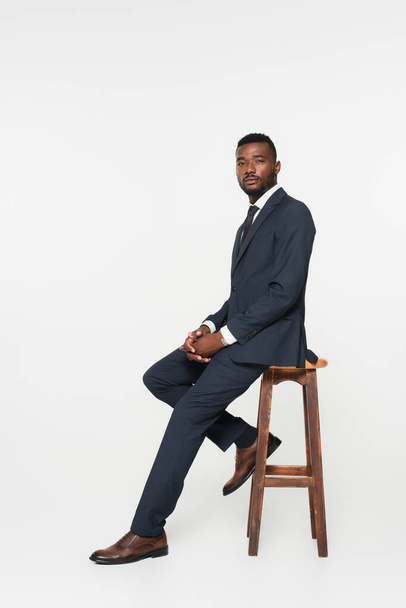 full length of african american businessman in suit sitting on wooden stool isolated on grey - Φωτογραφία, εικόνα