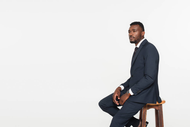 african american businessman in suit sitting on wooden stool isolated on grey - 写真・画像
