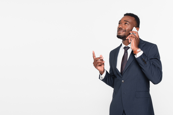 cheerful african american businessman in suit having phone conversation while pointing up with finger isolated on grey - Foto, Imagem