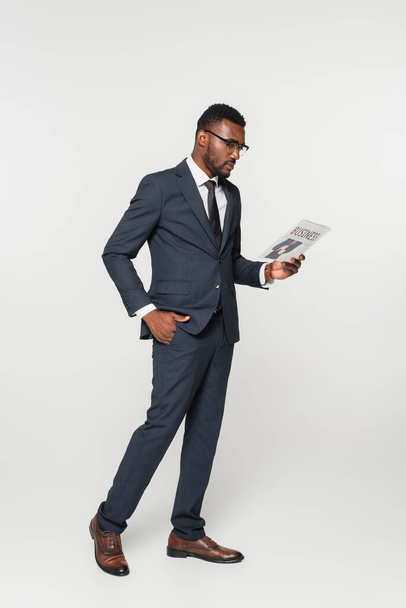 full length of african american man in eyeglasses reading business newspaper while standing with hand in pocket isolated on grey - Photo, Image