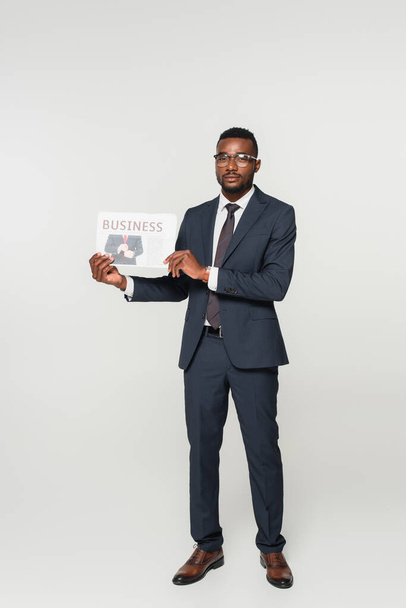 longitud completa de hombre afroamericano en gafas que muestran periódico de negocios aislado en gris - Foto, Imagen