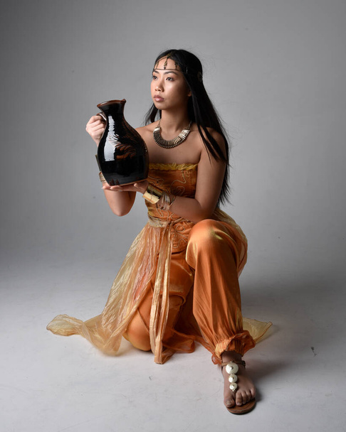 Ganztägiges Porträt einer hübschen jungen asiatischen Frau, die goldene arabische Gewänder trägt wie ein Genie, stehend in Pose mit einer Vase, isoliert auf Studiohintergrund. - Foto, Bild