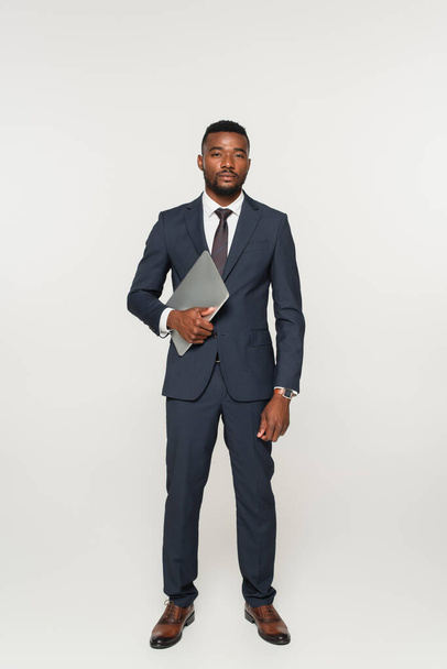 full length of african american businessman in suit holding laptop isolated on grey - Φωτογραφία, εικόνα