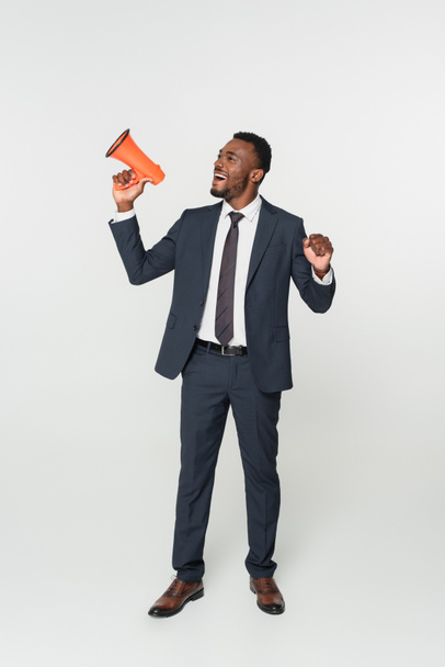 Ολόκληρο το μήκος του ενθουσιασμένου Αφροαμερικανού επιχειρηματία με κοστούμι ουρλιάζοντας σε μεγάφωνο απομονωμένο σε γκρι - Φωτογραφία, εικόνα