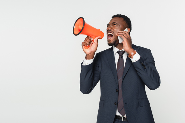 hombre de negocios afroamericano en traje gritando en megáfono y hablando en teléfono inteligente aislado en gris - Foto, Imagen