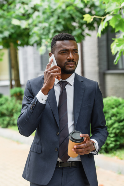 bebaarde Afro-Amerikaanse zakenman in pak met papieren beker en praten op mobiele telefoon  - Foto, afbeelding