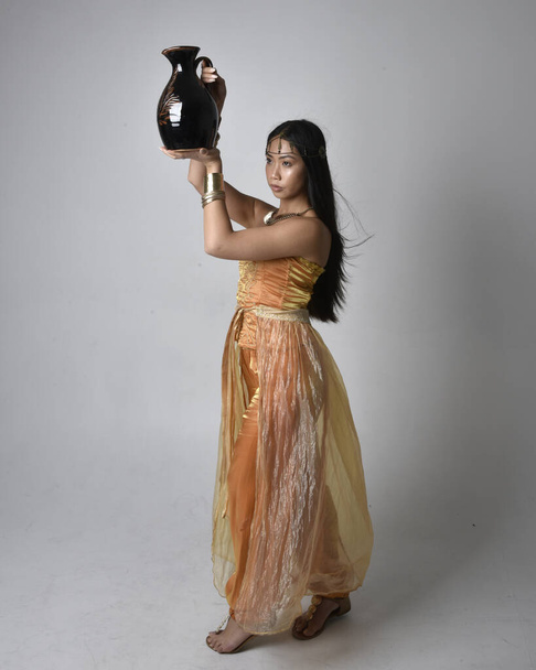 Full length portrait of pretty young asian woman wearing golden Arabian robes like a genie, standing pose holding a vase, isolated on studio background. - 写真・画像