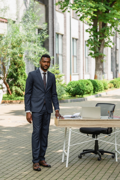 homme d'affaires afro-américain en costume debout près du bureau avec ordinateur portable à l'extérieur  - Photo, image