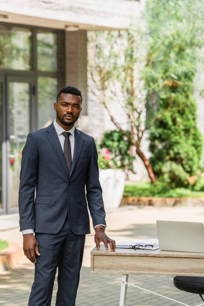 スーツ姿のアフリカ系アメリカ人ビジネスマンがノートパソコンを屋外に置いてデスクの近くに立ち  - 写真・画像