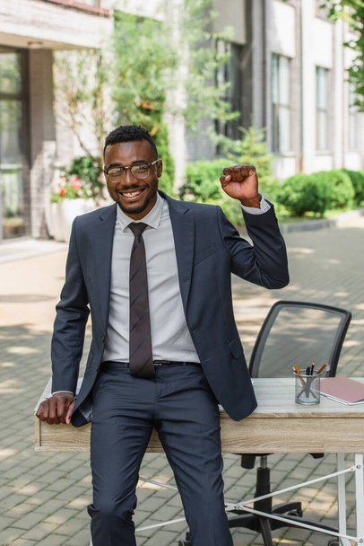 szczęśliwy Afrykański amerykański biznesmen w okularach z zaciśniętą pięścią opierającą się na biurku na zewnątrz  - Zdjęcie, obraz