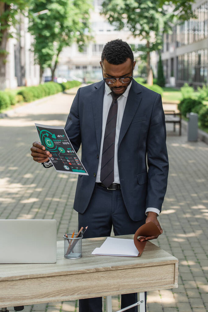Αφρικανός Αμερικανός επιχειρηματίας με γυαλιά ηλίου κοιτάζοντας το φάκελο κρατώντας εκτυπωμένα infographics  - Φωτογραφία, εικόνα