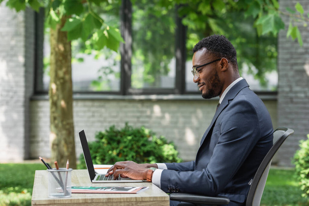 widok z boku wesoły afrykański amerykański biznesmen w okularach za pomocą laptopa  - Zdjęcie, obraz