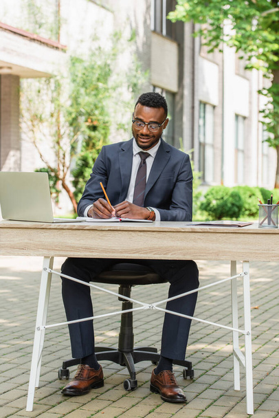 homme d'affaires afro-américain joyeux dans des lunettes écriture dans un ordinateur portable près de portable  - Photo, image