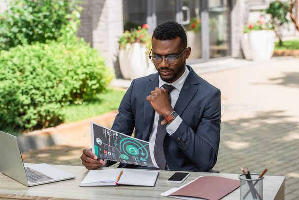 bearded african american businessman in eyeglasses looking at infographics near gadgets on desk - Photo, Image