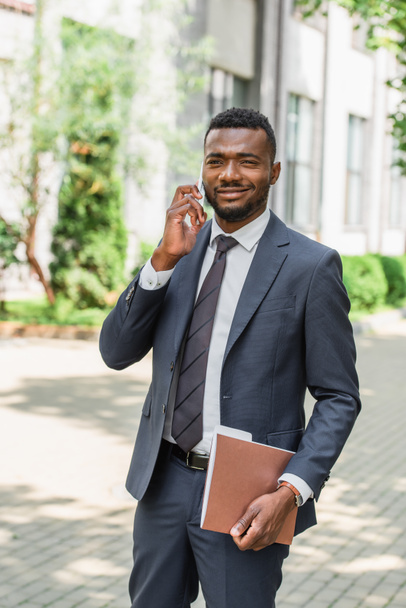 cheerful african american businessman holding folder and talking on smartphone outside - Foto, afbeelding