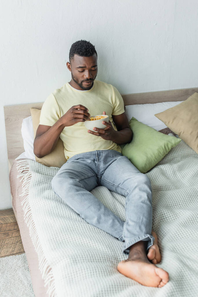 Çıplak ayaklı Afrikalı Amerikalı adam yatak odasında dinlenirken kasedeki mısır gevreklerine bakıyor. - Fotoğraf, Görsel