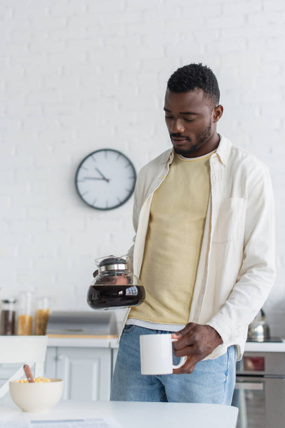 Afrikanisch-amerikanischer Mann hält Kaffeekanne neben Tasse in Küche - Foto, Bild