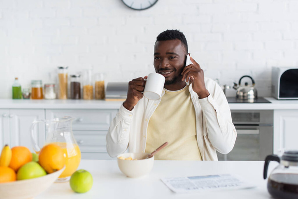 幸せなアフリカ系アメリカ人男性がコーヒーを飲みながらキッチンでスマホで話しています  - 写真・画像