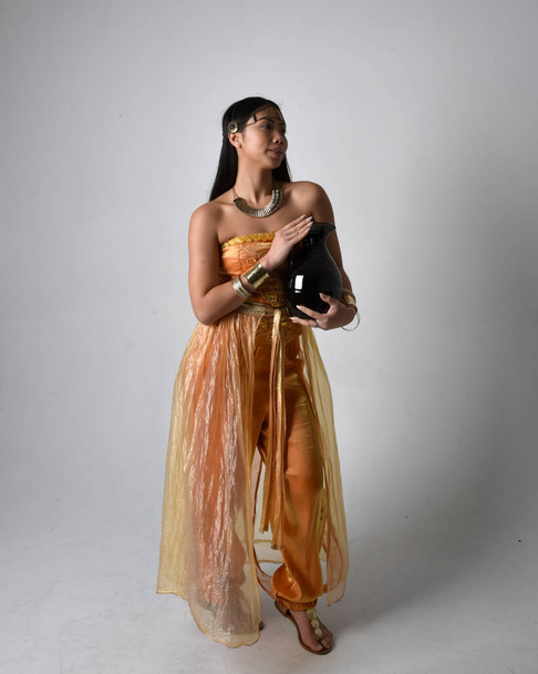 Retrato de larga duración de una hermosa joven asiática vestida con túnicas árabes doradas como un genio, de pie posando sosteniendo un jarrón, aislado en el fondo del estudio. - Foto, Imagen