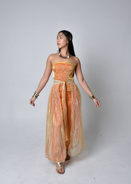 Ganztägiges Porträt einer hübschen jungen Asiatin, die goldene arabische Roben trägt wie ein Genie, mit dem Rücken zur Kamera posiert, isoliert auf Studiohintergrund. - Foto, Bild