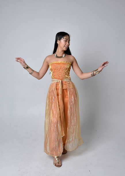 Full length portrait of pretty young asian woman wearing golden Arabian robes like a genie, standing pose  with back to the camera, isolated on studio background. - Zdjęcie, obraz