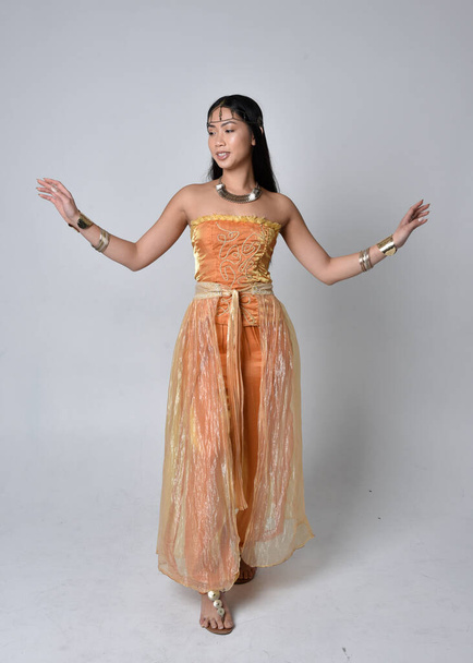 Full length portrait of pretty young asian woman wearing golden Arabian robes like a genie, standing pose  with back to the camera, isolated on studio background. - Foto, immagini