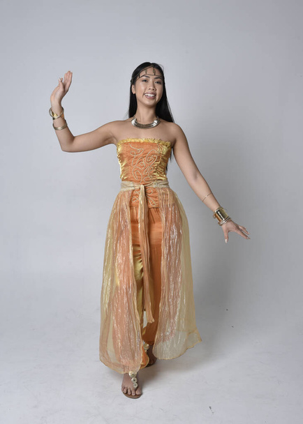 Full length portrait of pretty young asian woman wearing golden Arabian robes like a genie, standing pose  with back to the camera, isolated on studio background. - Photo, Image