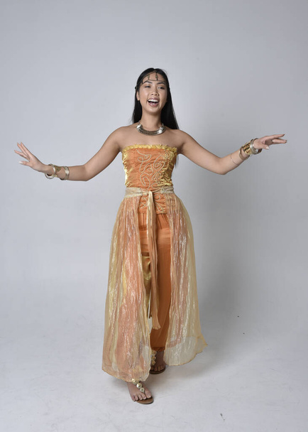 Full length portrait of pretty young asian woman wearing golden Arabian robes like a genie, standing pose  with back to the camera, isolated on studio background. - Foto, Imagem