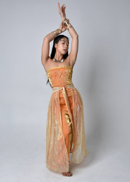 Retrato de comprimento total de mulher asiática muito jovem vestindo vestes árabes douradas como um gênio, em pé posar com volta para a câmera, isolado no fundo do estúdio. - Foto, Imagem