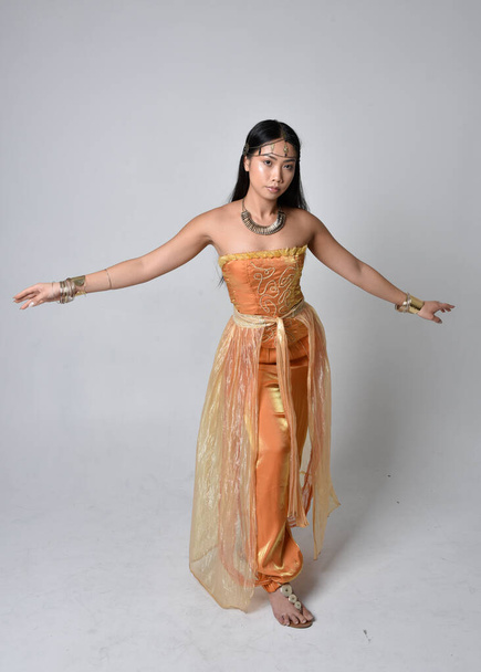 Full length portrait of pretty young asian woman wearing golden Arabian robes like a genie, standing pose  with back to the camera, isolated on studio background. - Photo, Image