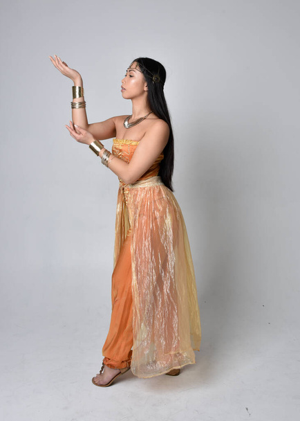 Full length portrait of pretty young asian woman wearing golden Arabian robes like a genie, standing pose  with back to the camera, isolated on studio background. - Foto, imagen