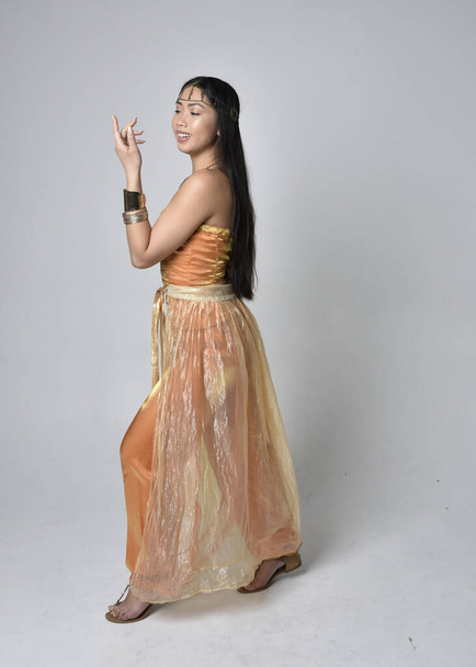 Full length portrait of pretty young asian woman wearing golden Arabian robes like a genie, standing pose  with back to the camera, isolated on studio background. - Foto, Bild