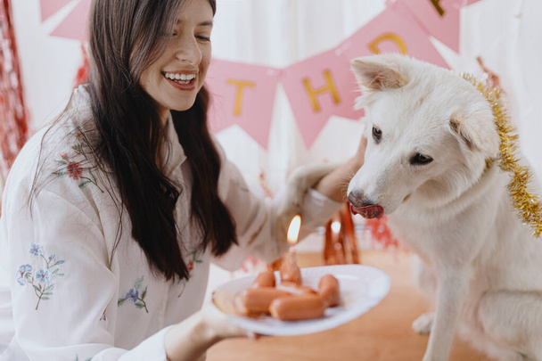 Une fête d'anniversaire de chien. Joyeux jeune femme et chien affamé célébrant l'anniversaire avec du gâteau à la saucisse et une bougie sur fond de guirlande rose. Adorable chien berger suisse blanc premier anniversaire - Photo, image