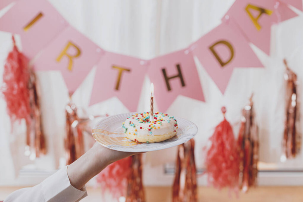 Νόστιμο ντόνατ γενεθλίων με κερί στο χέρι σε φόντο ροζ γιρλάντα και διακοσμήσεις στο εορταστικό δωμάτιο. Γιορτάζουμε τα γενέθλια. Πολύχρωμο ντόνατ με τρούφα και ροζ χρυσό κερί - Φωτογραφία, εικόνα