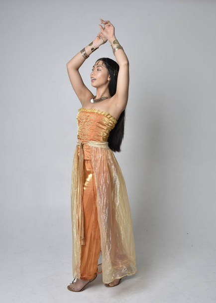 Retrato de larga duración de una hermosa joven asiática vestida con túnicas árabes doradas como un genio, posando de pie con la espalda a la cámara, aislada en el fondo del estudio. - Foto, Imagen