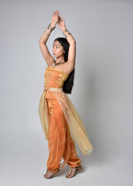 Full length portrait of pretty young asian woman wearing golden Arabian robes like a genie, standing pose  with back to the camera, isolated on studio background. - Photo, image