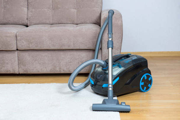 A new vacuum cleaner is standing next to the sofa, ready to start cleaning the room. - Zdjęcie, obraz