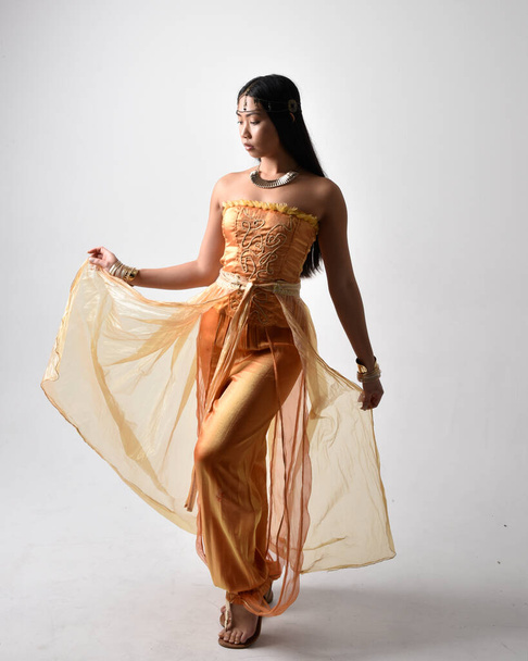 Retrato de larga duración de una hermosa joven asiática vestida con túnicas árabes doradas como un genio, posando de pie con la espalda a la cámara, aislada en el fondo del estudio. - Foto, Imagen