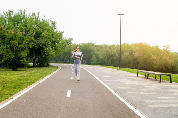 Woman dressed leggings and top running asphalt road summer park. Healthy lifestyle concept. Active and athletic female exercises oundoor - Photo, Image