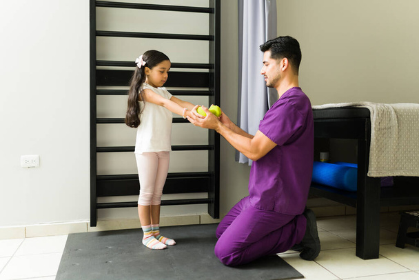 Pystyt siihen. Ammattimainen fysioterapeutti, joka kehottaa lapsipotilasta nostamaan käsipainon fyysiseen kuntoutukseensa - Valokuva, kuva